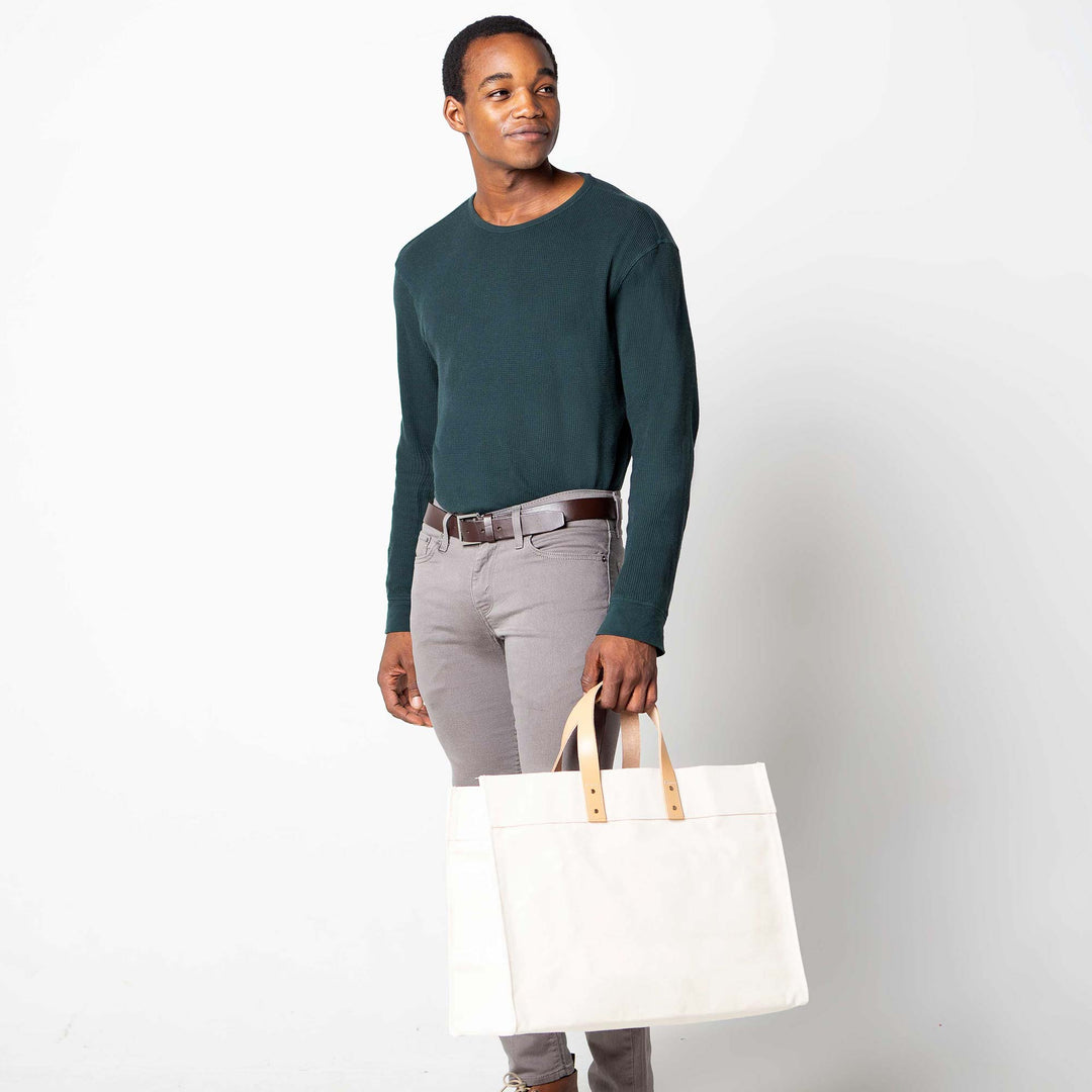 Man modeling dark teal long-sleeve shirt and gray pants while holding a blank white canvas utility tote bag with handles.