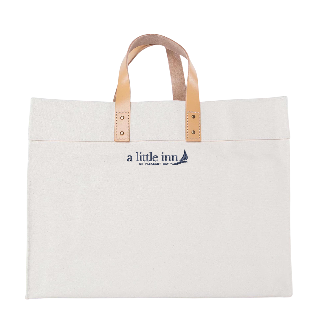 Natural canvas tote bag with tan leather handles and "a little inn" logo printed on front.