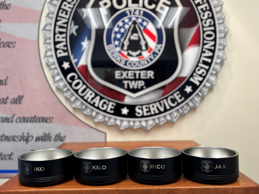 Four black stainless steel pet bowls with laser-engraved K9 unit call signs, displayed in front of an Exeter Police Department patch badge.