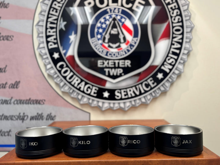 Four black stainless steel pet bowls with laser-engraved K9 unit call signs, displayed in front of an Exeter Police Department patch badge.
