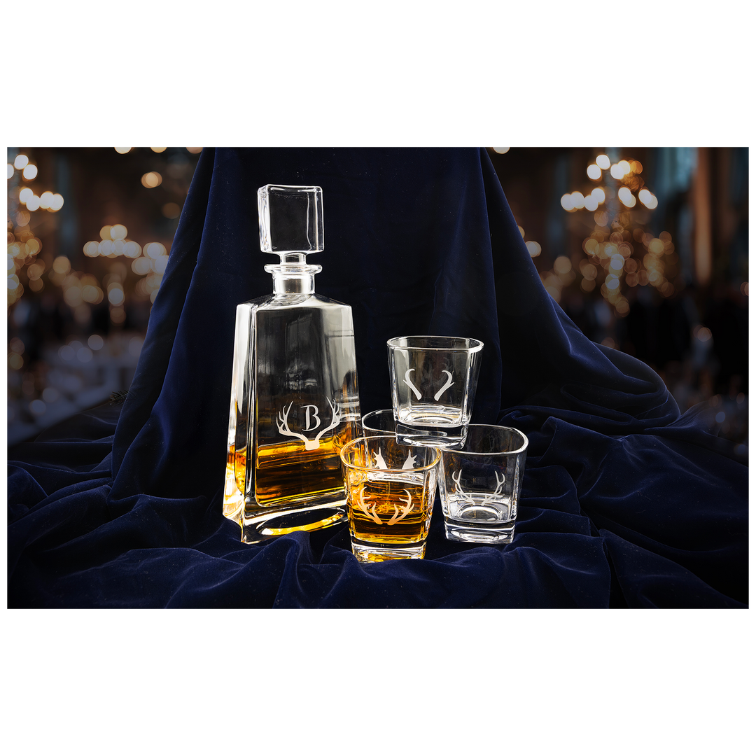 Glass decanter and three whiskey glasses with etched deer logo, filled with amber liquor, arranged on navy blue fabric.
