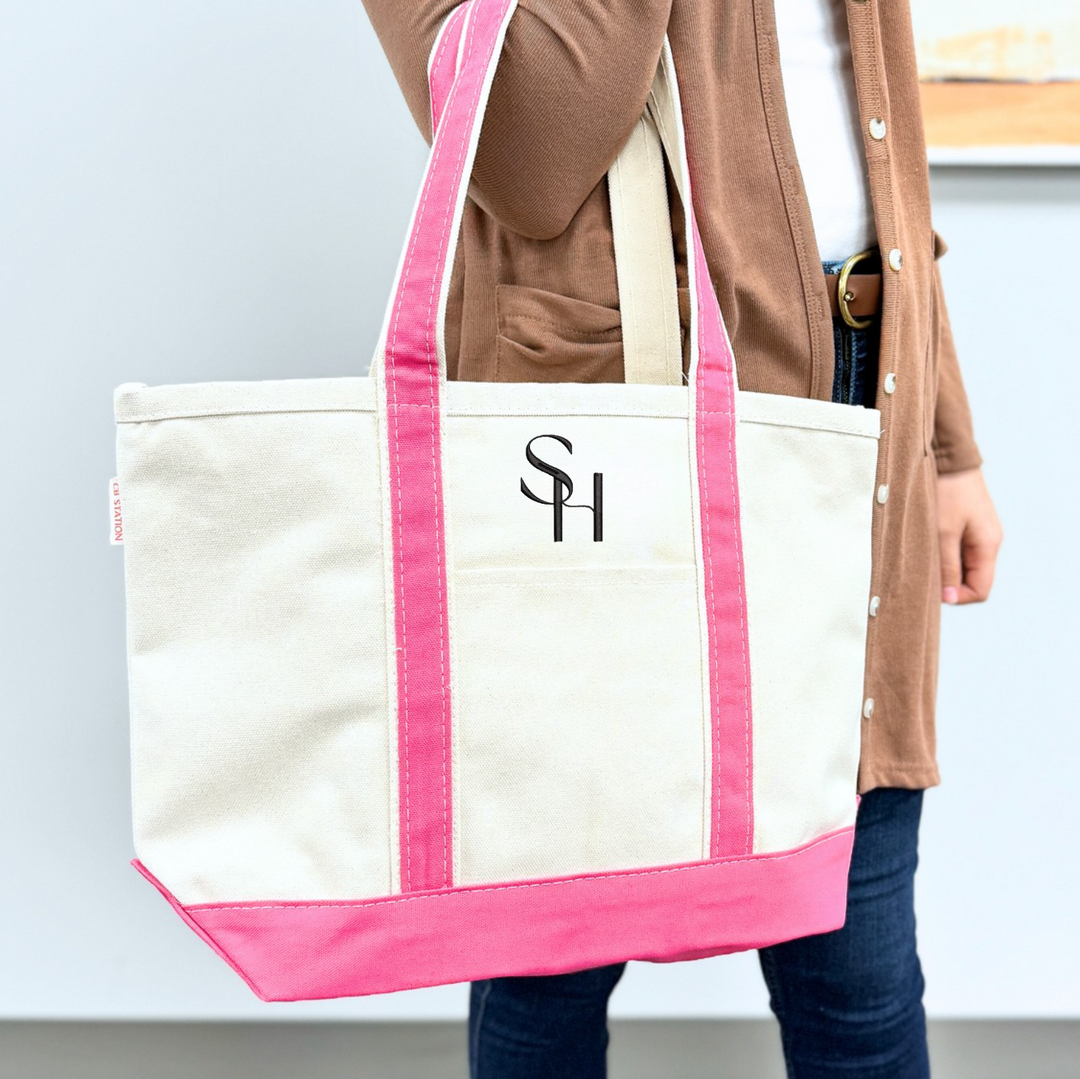 Cream canvas tote bag with hot pink handles and trim, featuring embroidered logo on front, held by person in brown cardigan.
