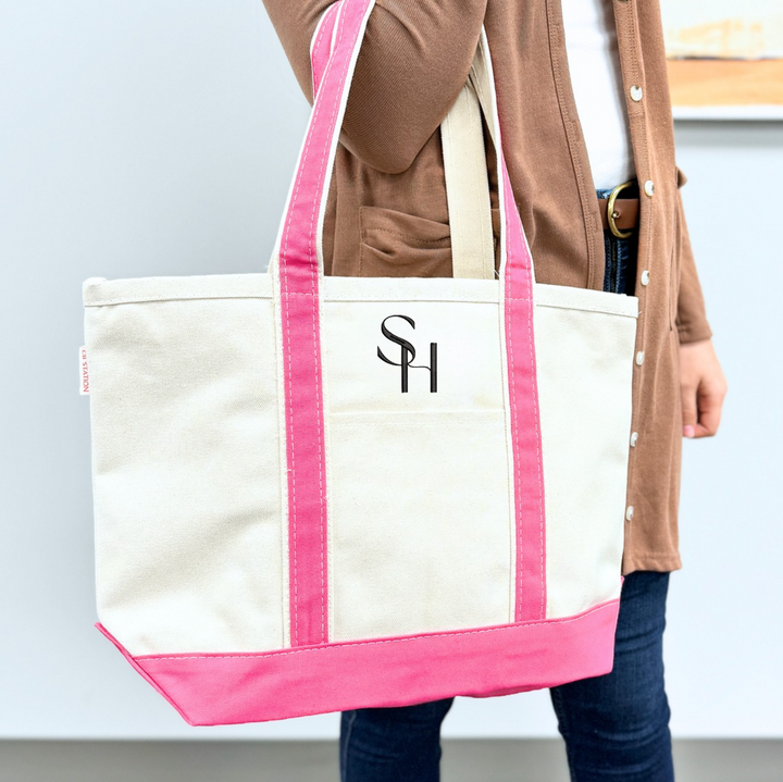 Cream canvas tote bag with hot pink handles and trim, featuring embroidered logo on front, held by person in brown cardigan.