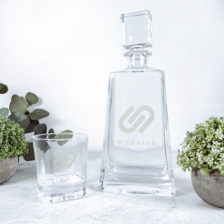 Clear glass tequila decanter with stopper and matching rocks glass, both etched with Modulus logo, displayed with potted greenery.