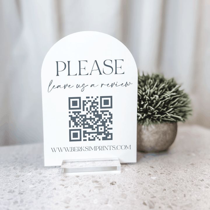 White arched acrylic sign with "PLEASE leave us a review" text and QR code linking to www.berksimprints.com, displayed on marble surface with potted plant.