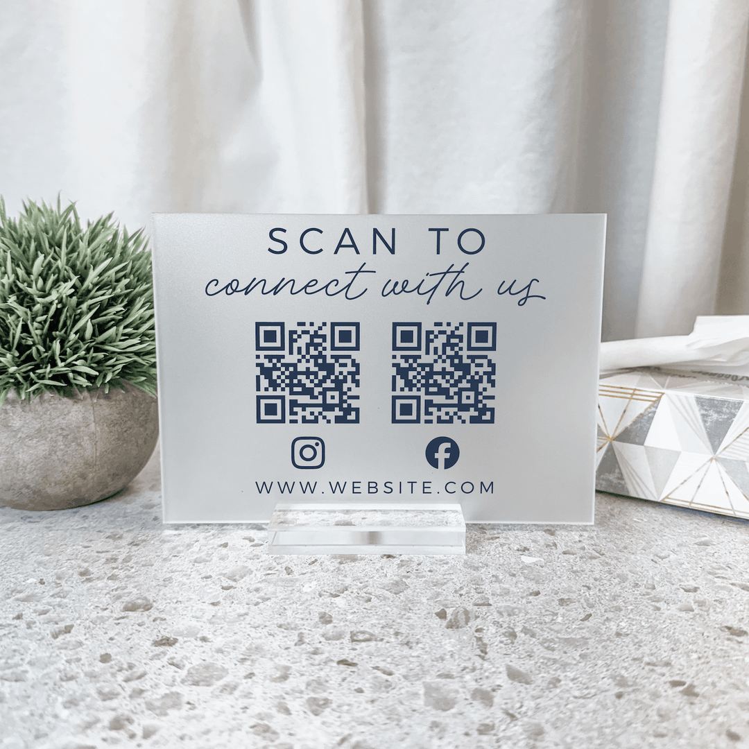 White acrylic sign with "Scan to Connect with us" text, two QR codes for Instagram and Facebook, and website URL displayed on terrazzo surface with potted plant.