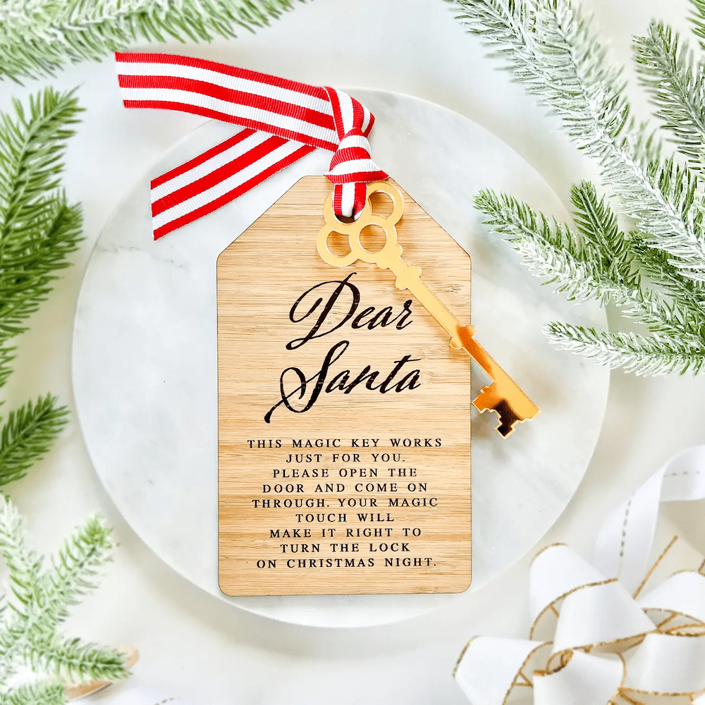 Bamboo "Dear Santa" door tag ornament with gold acrylic key and red striped ribbon bow, surrounded by evergreen sprigs on white background.
